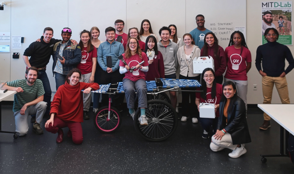 A group of MIT D-Lab students pose together in a classroom, standing and kneeling around a prototype stretcher mounted on bicycle wheels. Some students wear matching t-shirts with a lightbulb logo, and others hold small devices.